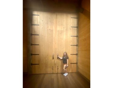 Lia stands in front of a large wooden door with her hand on the handle.